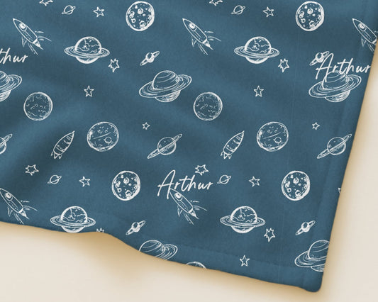 Fabric with space-themed pattern featuring planets, rockets, and stars on a blue background.