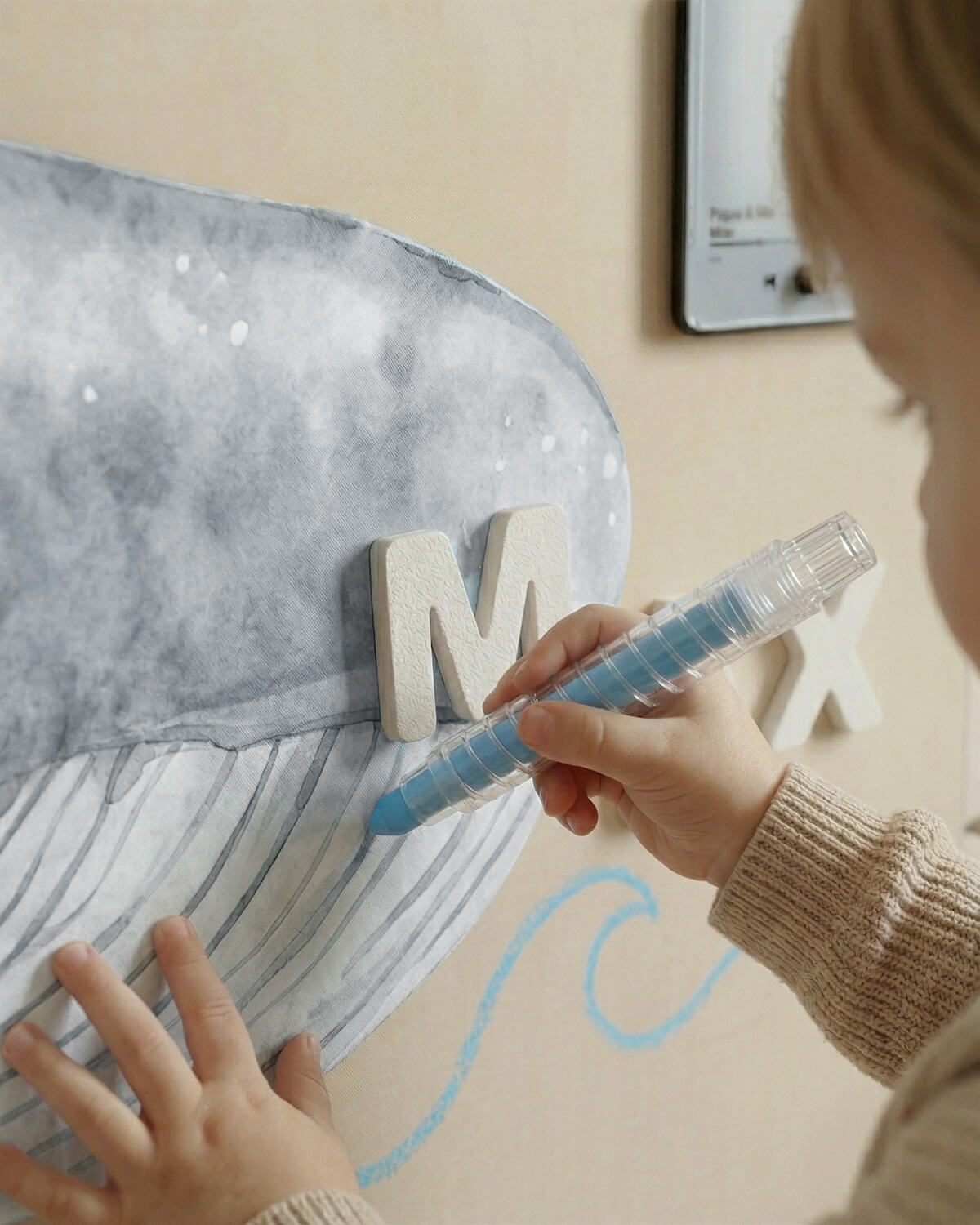 Child using a blue marker to draw on a large letter 'M' on a wall.