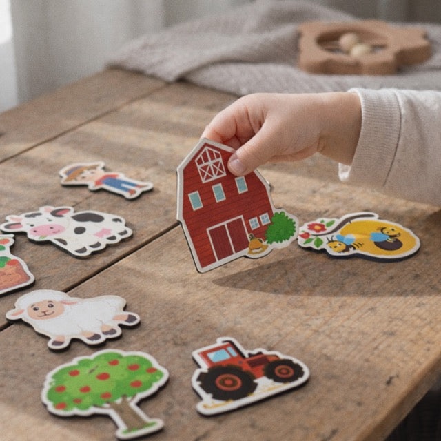 Child's hand holding a farm-themed magnets on a wooden surface with other magnets.
